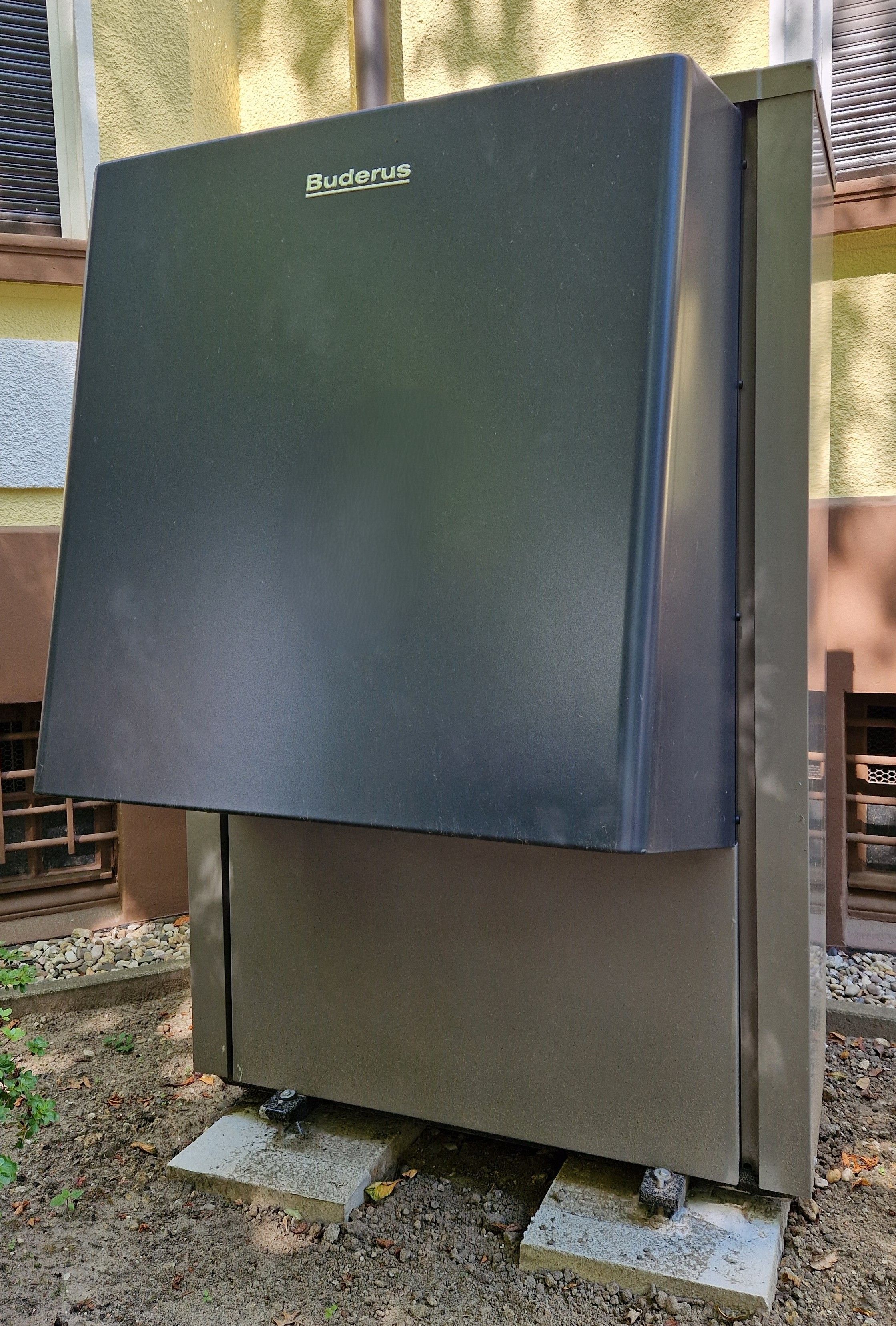Graues Buderus-Außengerät einer Wärmepumpe steht auf Betonblöcken im Schatten vor verputzter Hauswand zwischen Kies und Erde.