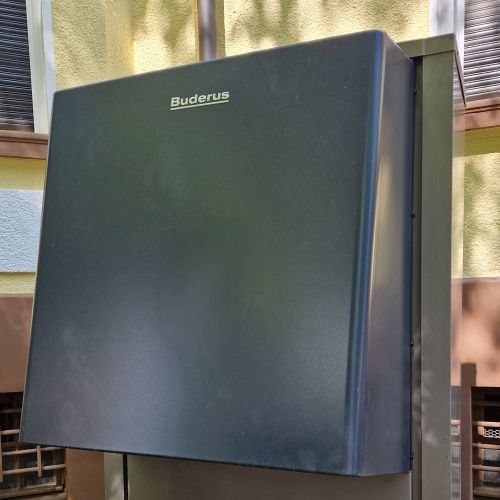 Graues Buderus-Außengerät einer Wärmepumpe steht auf Betonblöcken im Schatten vor verputzter Hauswand zwischen Kies und Erde.
