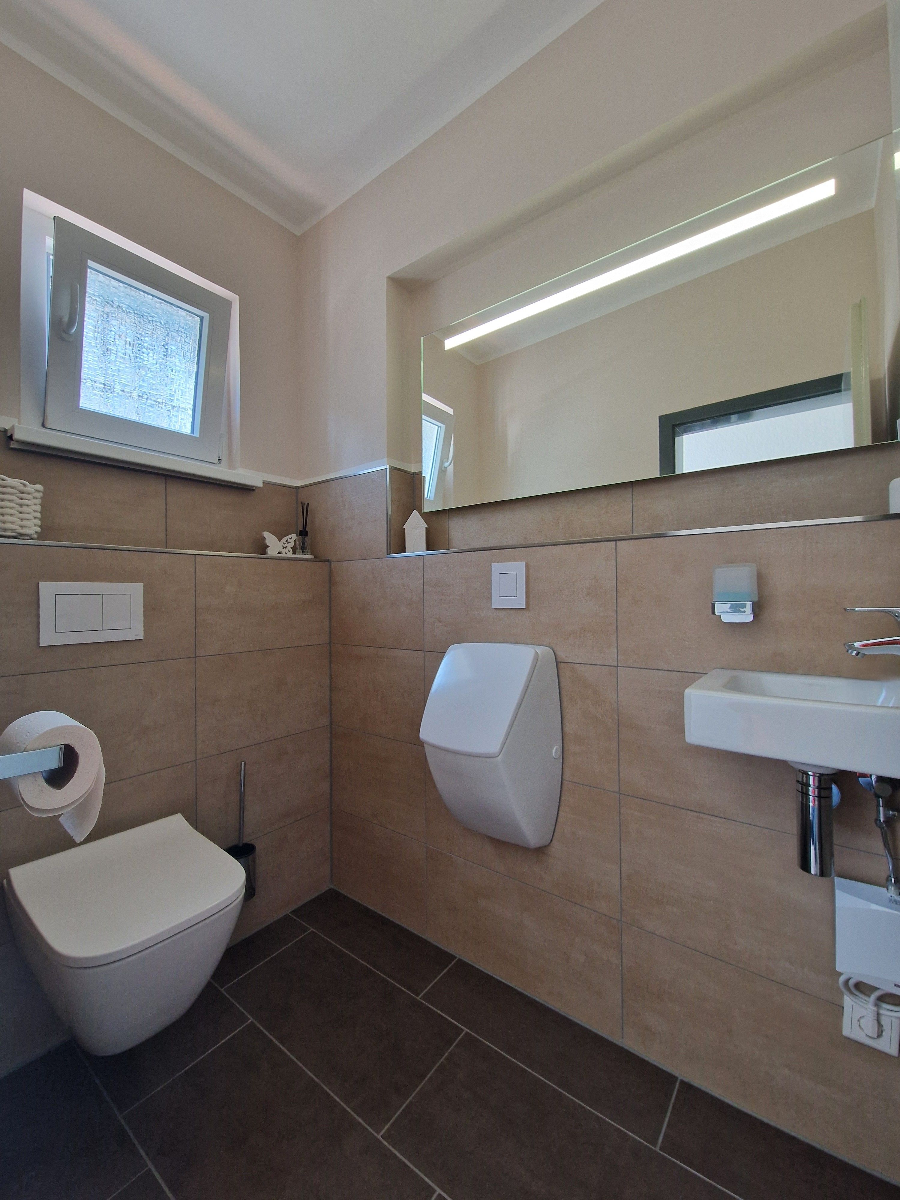 Zu sehen ist ein klassisch gefließtes Bad mit moderner Toilette, Pissoir und Waschbecken. Der Spiegel füllt die gesammte Wand.